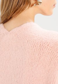 Light pink knitted sweater with a wide V-neck, showcasing a soft texture and loose fit. Hair is styled casually with a metallic earring.