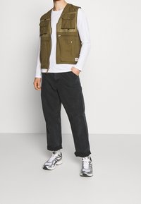 Man med olivgrön multifunktionell väst, vit långärmad skjorta, svarta byxor med uppvikta benslut, vita sneakers, står med ena handen i fickan.