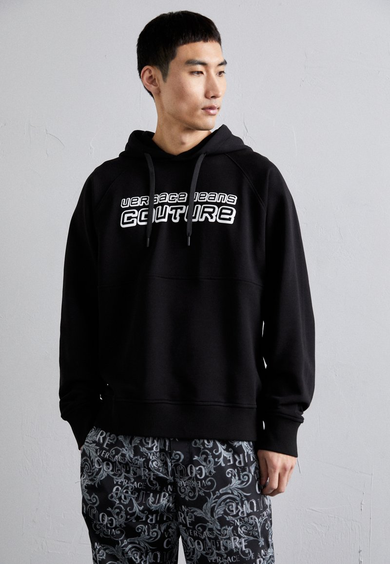 Black hooded sweatshirt with white "Versace Jeans Couture" logo. Features ribbed hem and cuffs, paired with patterned trousers in gray tones.