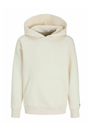 Teenagedreng iført en blå kasket, cremefarvet hættetrøje med frontlomme, sorte brede jeans og beige sko, stående og smilende på hvid baggrund.