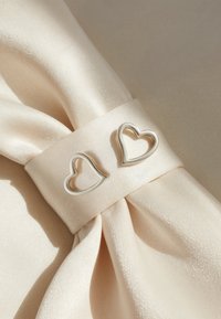 Silverfärgade hjärtformade metallaccenter på en krämfärgad satinband. Tyget har en slät, glänsande yta.