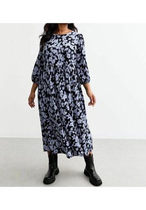 Femme portant une longue robe noire et grise à motifs avec des manches trois-quarts et des bottines noires, debout sur un sol clair.