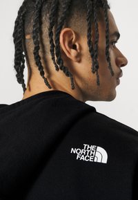 Felpa nera con un grande logo bianco "The North Face" sulla spalla sinistra. Il tessuto ha una finitura liscia e testurizzata.