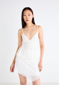 White dress with a pleated bodice and thin straps, featuring a layered peplum design and an asymmetric lace skirt. Smooth texture overall.
