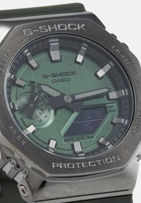 G-SHOCK watch with a stainless steel case, green dial, and digital display. Features silver hour markers and textured hands. Green strap.