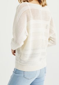 Valkoinen neulottu villapaita avoimella neulekuviolla, jossa on vaakaraidat ja ribbineuleinen helma. Yhdistetty vaaleansinisiin farkkuihin.