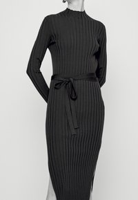 Femme portant une robe midi noire à manches longues et à côtes avec un col montant et une ceinture à nouer, se tenant devant un fond uni.