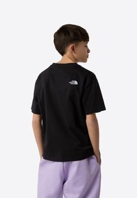 Black cotton t-shirt with a crew neck and short sleeves, featuring a small white logo on the upper back. Worn with light purple pants.
