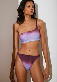 Ein Bikini-Oberteil mit einem Träger präsentiert einen Gradient von Hellblau zu Lila, mit einem geflochtenen Träger. Der passende Unterteil hat ein hochgeschnittenes Design.