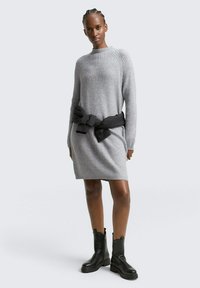 Robe en tricot rayé gris avec des manches longues et un col rond, accentuée par une ceinture en tissu noir nouée. Des bottines noires à la cheville complètent le look.