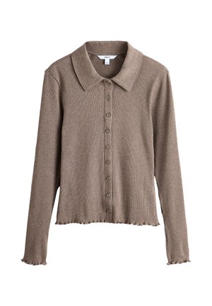 Jeune femme portant une veste en daim marron, une chemise grise boutonnée, un jean bleu et des mocassins marron, debout devant un fond beige uni.