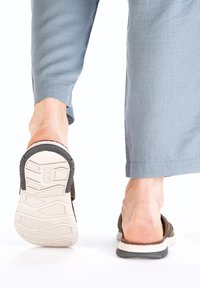 Brown slip-on shoes with a textured upper and a white, cushioned sole. The back is open, and the heel features a rubber grip. Light blue pants visible.