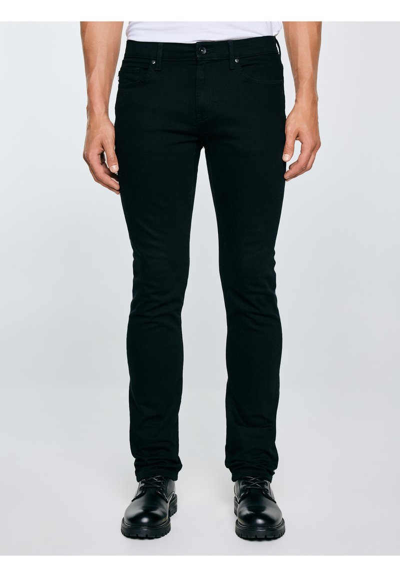 Férfi, aki fekete, vékony szabású farmert és fekete, fűzős csizmát visel, és egy egyszínű, világos háttér előtt áll.