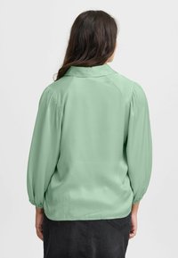 Kerge roheline, kraega blouse pikas, kogutud varrukatega ja veidi lõdva lõikega. Sile tekstuur ja lihtne, puhas disain.