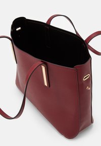 Otevřená bordó kožená taška přes rameno s dvěma rukojeťmi, černým interiérem a zlatými kovovými akcenty na bílém pozadí.