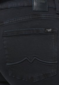 Közelkép sötét farmernadrágról, amely bemutatja a hátsó zsebet, jellegzetes ívelt varrással és egy kis ló logócímkével.