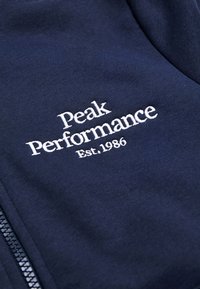 Felpa con zip di colore navy in cotone, con testo ricamato bianco "Peak Performance Est. 1986." Texture liscia con dettagli a coste.