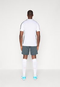 Camiseta atlética de mangas curtas brancas com detalhes laterais pretos, combinada com calções cinzentos. Meias brancas e chuteiras azul claro com detalhes em logotipo.