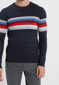 Pull en tricot bleu marine à manches longues, avec des rayures horizontales rouges, bleues, grises et blanches sur la poitrine. Coupe régulière.