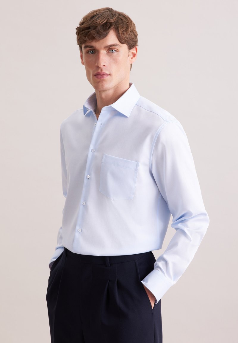 Jeune homme portant une chemise bleu clair à boutons et un pantalon noir, debout les mains dans les poches devant un fond uni.