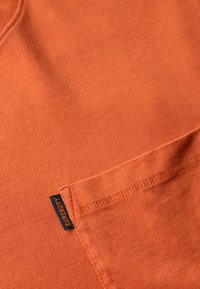 Terrakotta T-shirt lavet af blød bomuld. Har en brystlomme og et lille sort mærke med "Superdry." Glat, ensartet tekstur.