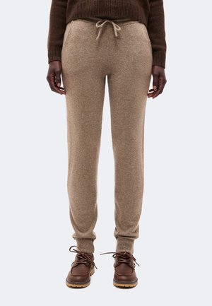 Personne portant un pantalon de jogging en maille beige avec taille à cordon, un pull marron et des bottes marron à lacets sur un fond clair uni.