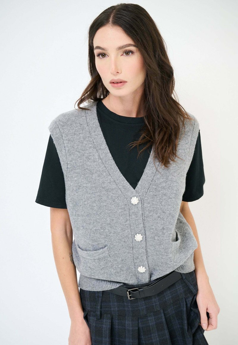 Gilet grigio lavorato a maglia con scollo a V, caratterizzato da quattro bottoni decorativi, due tasche frontali, indossato sopra una t-shirt nera, abbinato a una gonna a quadri.