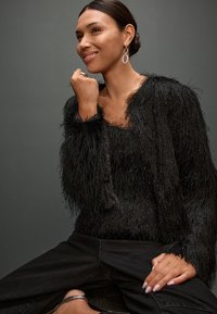 Top negro peludo con detalles de flecos y cuello de satén, combinado con pantalones de mezclilla negros. La modelo lleva pendientes ovalados plateados y un anillo.