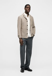 Muž v béžovom zapínacom blejzri, bielej košeli, sivých nohaviciach a čiernych elegantných topánkach stojí na bielom pozadí.