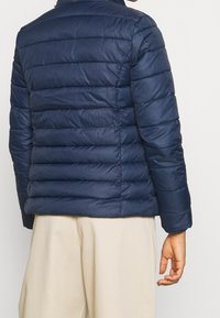 Tmavě modrá prošívaná bunda s horizontálním prošíváním, lehká tkanina, stojatý límec a elastické manžety. Nositelná s kalhotami světlé barvy.