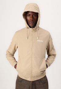 Beige huva jacka med frontdragkedja, elastiska ärmslut och sidofickor. "JACK&JONES" logo i vitt, slät yta och avslappnad design.