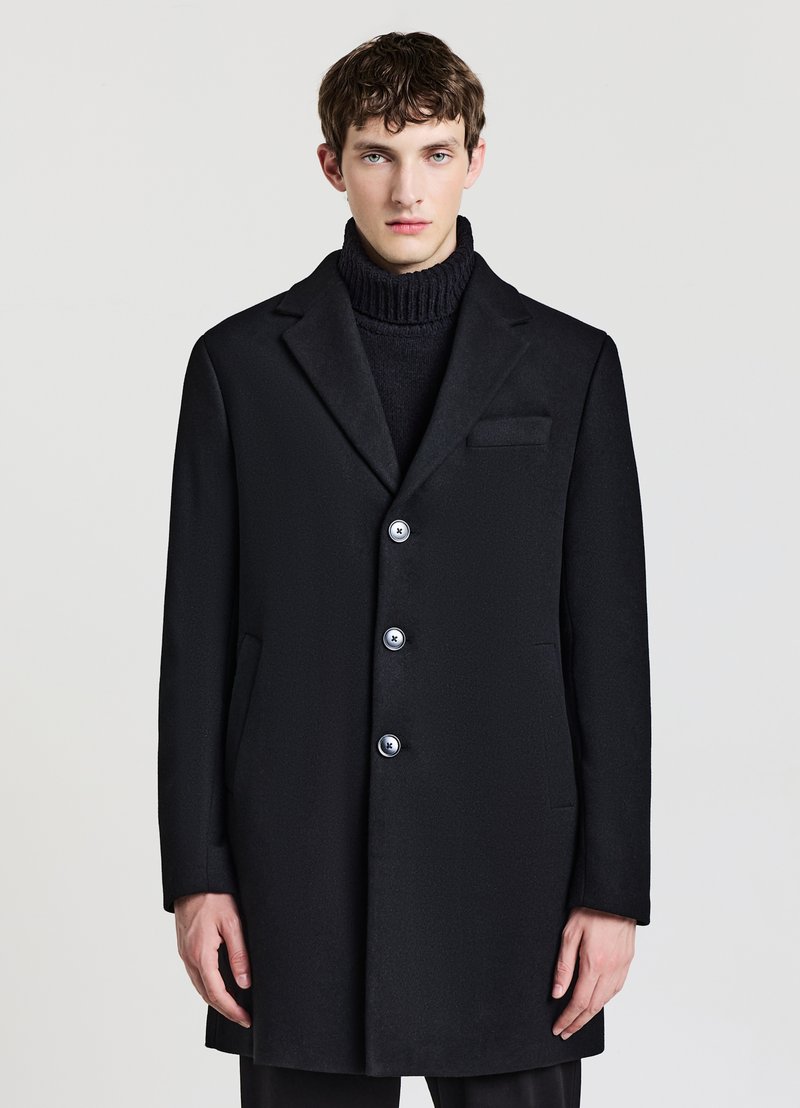 Schwarzer Wollmantel mit Reverskragen, fünf Knöpfen zum Schließen, Seitentaschen und schlanker Passform. Getragen über einem strukturierten schwarzen Rollkragenpullover.