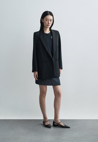 Μαύρο υπερμεγέθες σακάκι με αυλακωτούς πέλματος, συνδυασμένο με εφαρμοστό μαύρο τοπ και σκούρα τζιν φούστα. Το σύνολο ολοκληρώνεται με μαύρες μυτερές γόβες.