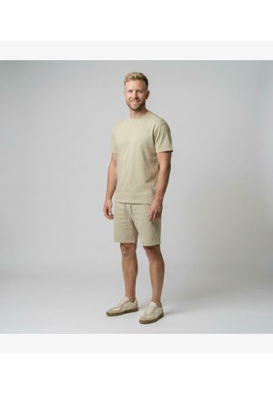 Man staat glimlachend, draagt een beige shirt met korte mouwen, bijpassende korte broek en beige sneakers tegen een effen lichtgrijze achtergrond.