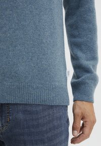 Pull en tricot bleu avec des poignets et un ourlet côtelés, présentant une texture subtile. Étiquette visible sur le côté gauche. Porté avec un jean en denim foncé.
