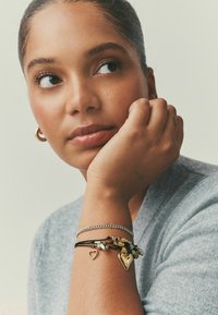 Sølv kædearmbånd og læder snorarmbåndstak med hjerteformede charms i guld og sølv, båret på en solbrun arm.