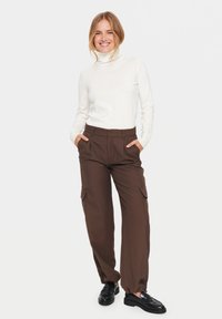 Witte coltrui, slim fit; bruine cargobroek met zijzakken; zwarte instapschoenen; staande pose tegen een effen witte achtergrond.