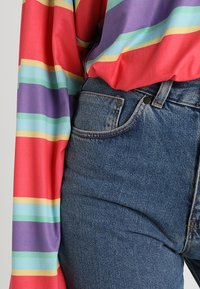 Monivärinen pitkähihaistoppi, jossa on punaisia, violetteja ja teal-värisiä raitoja, yhdistettynä korkeavyötäröisiin sinisiin denimgn haalarisiin, joissa on perinteinen taskusuunnittelu.