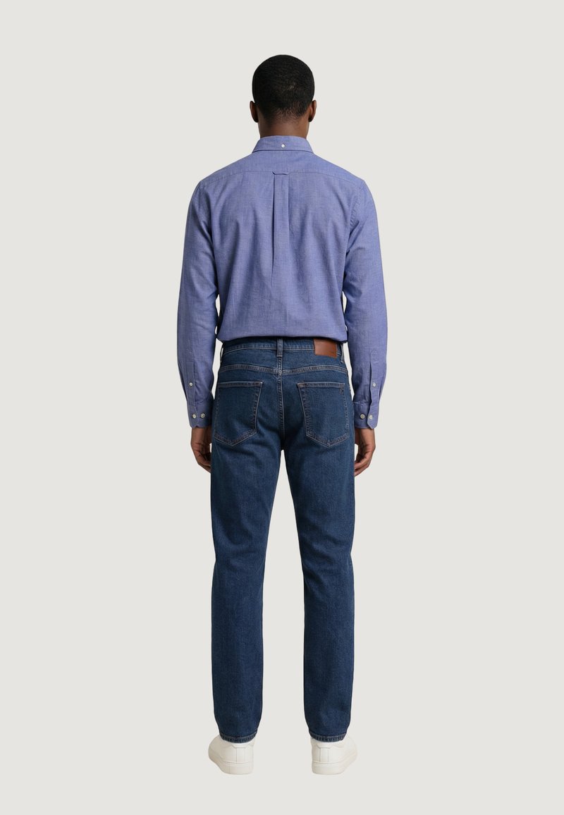 Homme portant une chemise bleue à manches longues boutonnée, rentrée dans un jean bleu foncé, avec des baskets blanches, debout, dos tourné, sur un fond clair.