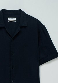 Camisa de algodón de manga corta en azul marino oscuro con cuello tipo camp, cierre de botones y un logo sutil en el lado izquierdo del pecho.