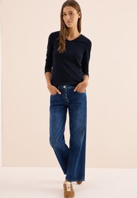 Marinefarbener Langarm-V-Ausschnitt-Oberteil kombiniert mit weit geschnittenen blauen Denim-Jeans. Das Outfit wird durch hellbraune Sneakers ergänzt. Einfaches Design, lässiger Look.