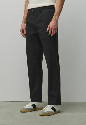 Mężczyzna ubrany w czarne spodnie o prostych nogawkach i białe sneakersy z szarymi i czarnymi akcentami, stojący na szarej podłodze na tle gładkiej ściany.