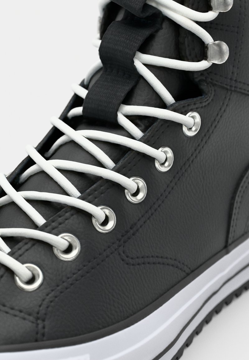 Baskets en cuir noir avec des lacets blancs, des œillets en métal et une surface texturée. La semelle est blanche avec un motif de bande de roulement noir.