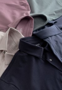 Fünf ordentlich gefaltete Poloshirts in gedämpftem Rosa, Beige, Navyblau, Hellblau und Salbeigrün mit sichtbaren Kragen und Knöpfen.