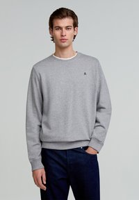 Grauer Sweatshirt aus Baumwolle mit Rundhalsausschnitt, gerippten Bündchen und Saum. Verfügt über ein kleines gesticktes Logo auf der Brust. Getragen zu dunkelblauen Jeans.