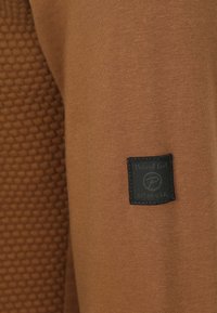 Bruine sweatshirt met een textuurpatroon aan de ene kant en een zwart label met logo aan de andere kant. Zacht materiaal, casual ontwerp.