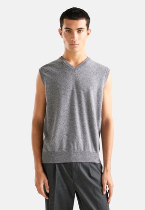 Suéter gris sin mangas con un diseño de escote en V, hecho de tejido de punto. Textura suave, dobladillo acanalado, estilo ajustado, combinado con pantalones oscuros de estampado.