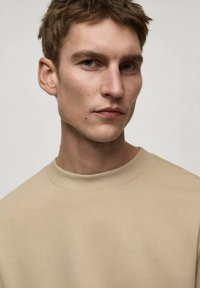 Beige mikina s kulatým výstřihem a žebrovanými manžetami. Hladký materiál, minimalistický design. Model s krátkými vlasy a neutrálním pozadím.