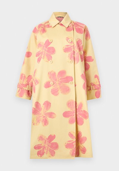 Long manteau trench jaune double boutonnage avec de grandes imprimés floraux roses délavés et des sangles à boucle sur les manches.