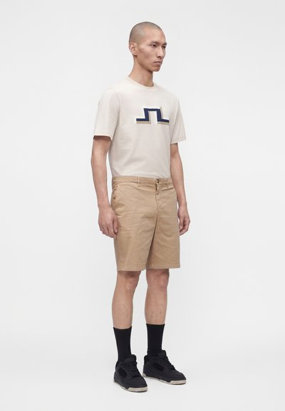 Homme debout portant un t-shirt graphique beige, un short beige à hauteur des genoux, des chaussettes noires montantes et des baskets noires sur fond blanc uni.